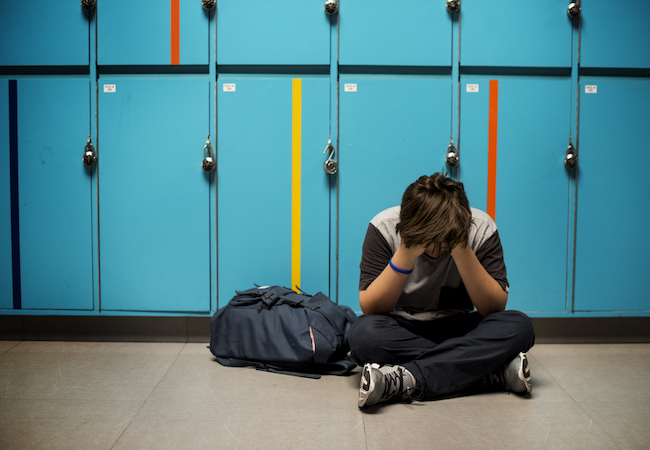 Student with head in hands sitting on the floor in front of blue lockers. 